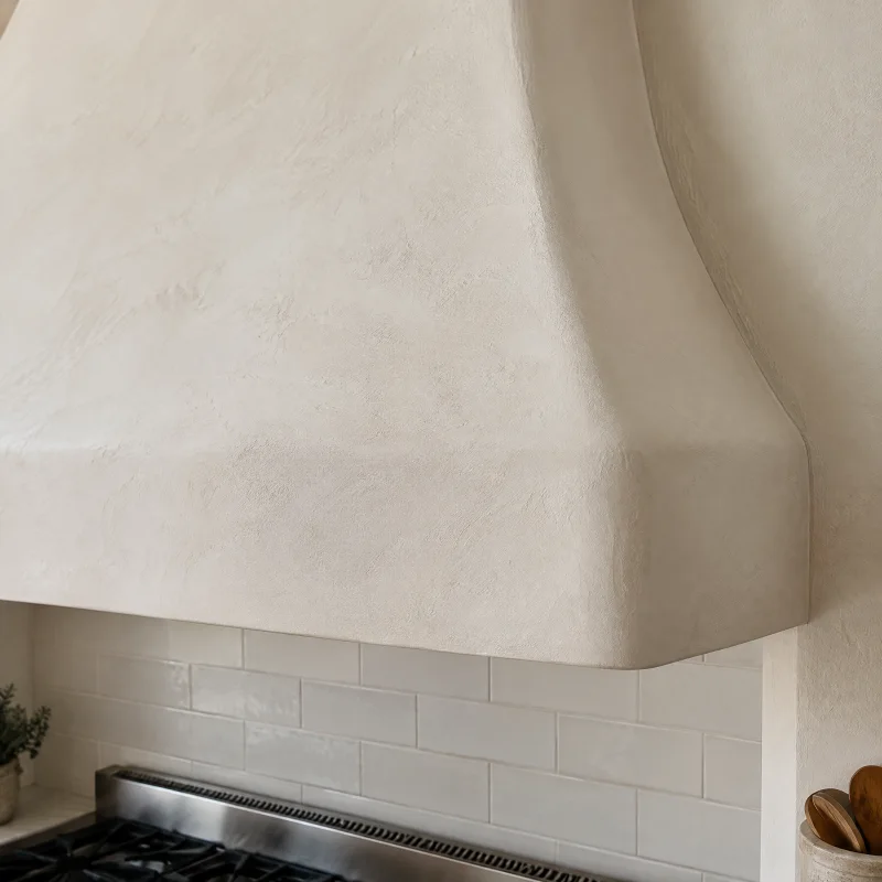 Close-up of a warm white plaster range hood with soft rounded edges and a subtle skim texture meeting a plaster wall